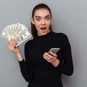 Surprised happy brunette woman in black clothes holding money and smartphone while looking at the camera over gray background