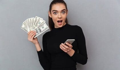 Surprised happy brunette woman in black clothes holding money and smartphone while looking at the camera over gray background