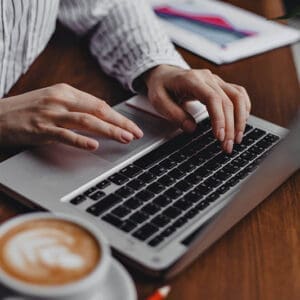A female hands press laptop keys while sitting at a wooden table with a white cappuccino cup, and she is working to start a blog.